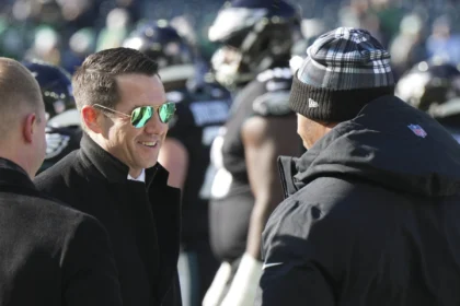 Ein Mann mit schwarzem Mantel und Sonnenbrille lächelt, während er mit einer anderen Person, die eine Wintermütze trägt, auf einem Footballfeld in New York spricht. Im Hintergrund sind Spieler zu sehen, die möglicherweise über die Verlängerung des Vertrags von GM Joe Schoen diskutieren. Alt-Text wurde KI-generiert.