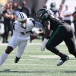 Ein Footballspieler in einer weißen Uniform blockt einen Seahawks-Spieler in einer schwarzen Uniform während eines Spiels auf einem sonnigen Feld. Alt-Text wurde KI-generiert.