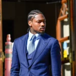Stefon Diggs - Ein Mann in blauem Anzug und Krawatte steht im Freien in der Nähe eines Gebäudeeingangs und schaut nach rechts, was an Stefon Diggs' souveräne Präsenz abseits des Spielfelds erinnert. Alt-Text wurde KI-generiert.