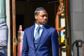 Stefon Diggs - Ein Mann in blauem Anzug und Krawatte steht im Freien in der Nähe eines Gebäudeeingangs und schaut nach rechts, was an Stefon Diggs' souveräne Präsenz abseits des Spielfelds erinnert. Alt-Text wurde KI-generiert.