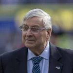 Ein älterer Mann mit grauen Haaren und Brille trägt einen Anzug, ein blaues Hemd und eine Krawatte mit Polka-Dots. Er steht im Freien vor einem unscharfen Hintergrund und strahlt dabei einen dezenten Stolz auf die Buffalo Bills aus. Alt-Text wurde KI-generiert.