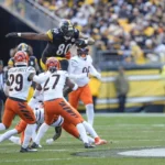Ein Spieler der Pittsburgh Steelers, inspiriert von Darnell Washington kürzlicher Vertragsverlängerung, springt während eines Footballspiels an einem sonnigen Tag über zwei Verteidiger der Cincinnati Bengals. Alt-Text wurde KI-generiert.