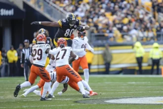 Ein Spieler der Pittsburgh Steelers, inspiriert von Darnell Washington kürzlicher Vertragsverlängerung, springt während eines Footballspiels an einem sonnigen Tag über zwei Verteidiger der Cincinnati Bengals. Alt-Text wurde KI-generiert.