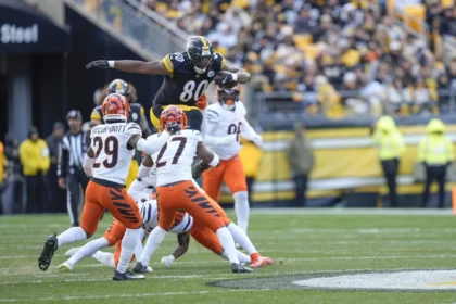 Ein Spieler der Pittsburgh Steelers, inspiriert von Darnell Washington kürzlicher Vertragsverlängerung, springt während eines Footballspiels an einem sonnigen Tag über zwei Verteidiger der Cincinnati Bengals. Alt-Text wurde KI-generiert.