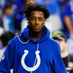 Ein Mann, der einen blauen Indianapolis-Colts-Hoodie und Kopfhörer trägt, steht in einem Stadion, im Hintergrund sind verschwommene Zuschauer zu sehen; möglicherweise streamt er inmitten der Menge Internet-Highlights von Sauce Gardner. Alt-Text wurde KI-generiert.