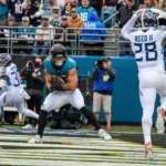 Brenton Strange, Tight End der Jaguars, im blaugrünen Trikot, fängt einen Pass in der Endzone, während ein Verteidiger in Weiß die Arme hebt; Zuschauer und Fotografen beobachten das Geschehen von der Seitenlinie aus. Alt-Text wurde KI-generiert.