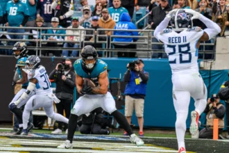 Brenton Strange, Tight End der Jaguars, im blaugrünen Trikot, fängt einen Pass in der Endzone, während ein Verteidiger in Weiß die Arme hebt; Zuschauer und Fotografen beobachten das Geschehen von der Seitenlinie aus. Alt-Text wurde KI-generiert.