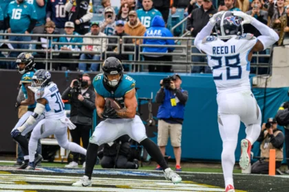 Brenton Strange, Tight End der Jaguars, im blaugrünen Trikot, fängt einen Pass in der Endzone, während ein Verteidiger in Weiß die Arme hebt; Zuschauer und Fotografen beobachten das Geschehen von der Seitenlinie aus. Alt-Text wurde KI-generiert.