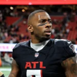 Ein Footballspieler im Trikot der Falcons steht auf dem Spielfeld, blickt zur Seite, im Hintergrund sind das Stadion und die Zuschauer zu sehen – ganz ähnlich wie Kyle Pitts, nachdem er seinen Dreijahresvertrag unterzeichnet hatte. Alt-Text wurde KI-generiert.
