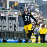 Ein Football-Spieler in einem schwarz-gelben Trikot läuft auf das Spielfeld und zeigt nach oben. Im Hintergrund ist ein "Steelers"-Schild zu sehen, und die Fans aus Pittsburgh schauen zu - sie feiern wie Nick Herbig nach seinem Vertrag mit den Steelers. Alt-Text wurde KI-generiert.