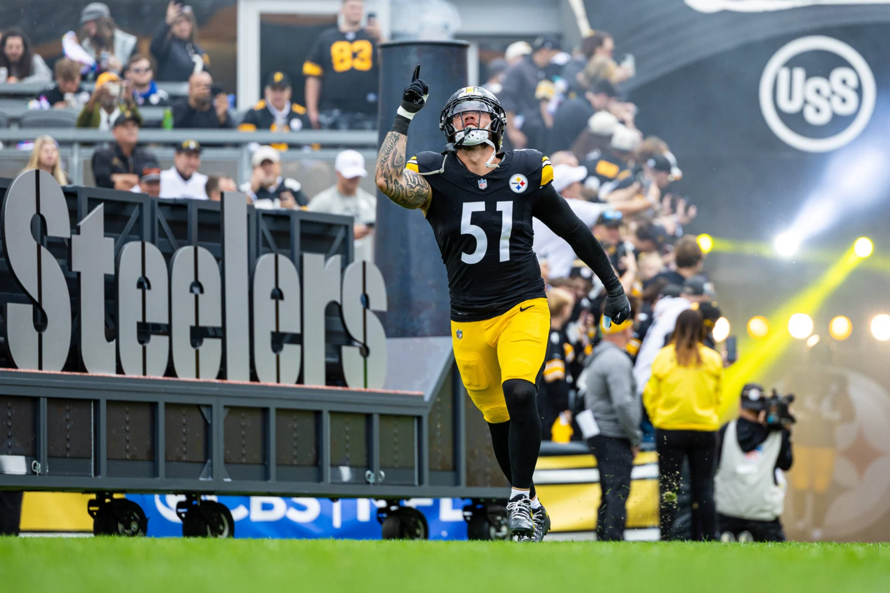 Ein Football-Spieler in einem schwarz-gelben Trikot läuft auf das Spielfeld und zeigt nach oben. Im Hintergrund ist ein "Steelers"-Schild zu sehen, und die Fans aus Pittsburgh schauen zu - sie feiern wie Nick Herbig nach seinem Vertrag mit den Steelers. Alt-Text wurde KI-generiert.
