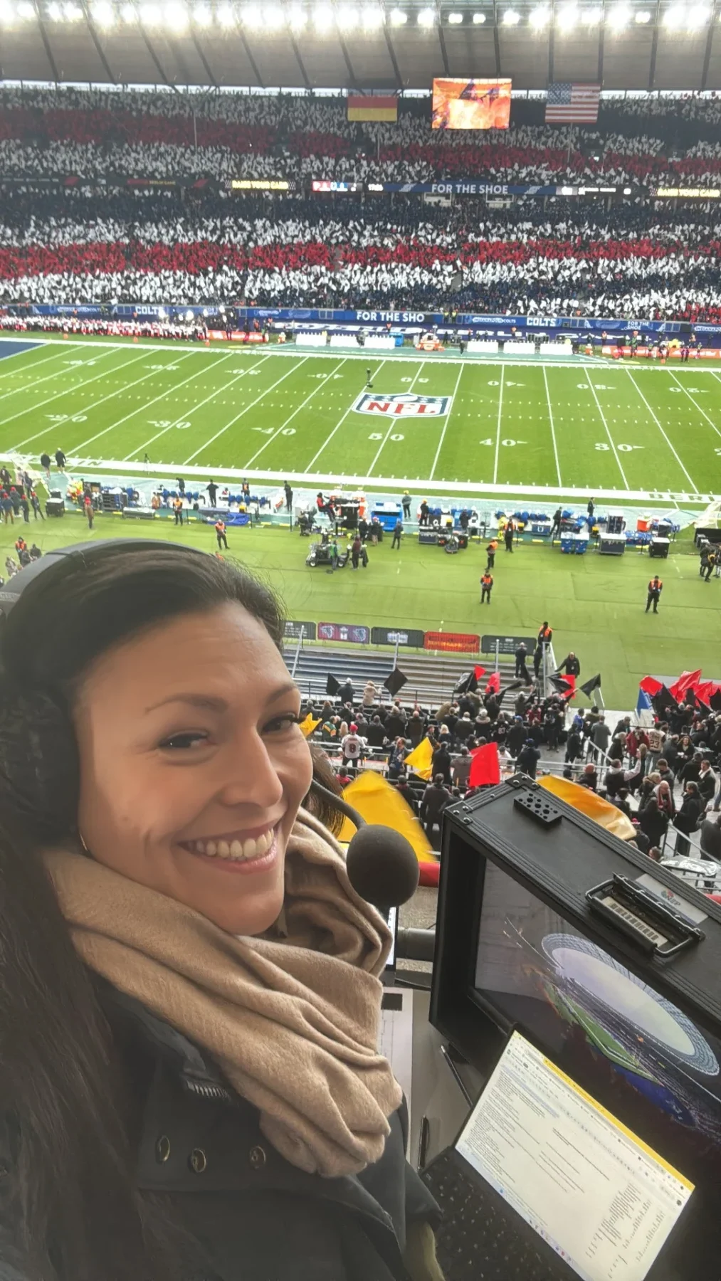 Eine Frau mit Kopfhörern und Schal, die der deutschen NFL-Expertin Nadine Nurasyid ähnelt, lächelt während eines Spiels für ein Selfie in der Pressetribüne eines Stadions mit Blick auf das Spielfeld der Indianapolis Colts. Alt-Text wurde KI-generiert.