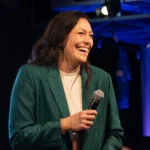 Eine Frau in einem blaugrünen Anzug, die NFL-Expertin Nadine Nurasyid, hält ein Mikrofon und ein Blatt Papier in der Hand und lächelt, während sie auf einer Bühne steht, im Hintergrund sind Trikots der Indianapolis Colts und anderer Sportmannschaften zu sehen. Alt-Text wurde KI-generiert.