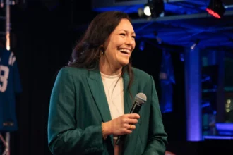 Eine Frau in einem blaugrünen Anzug, die NFL-Expertin Nadine Nurasyid, hält ein Mikrofon und ein Blatt Papier in der Hand und lächelt, während sie auf einer Bühne steht, im Hintergrund sind Trikots der Indianapolis Colts und anderer Sportmannschaften zu sehen. Alt-Text wurde KI-generiert.