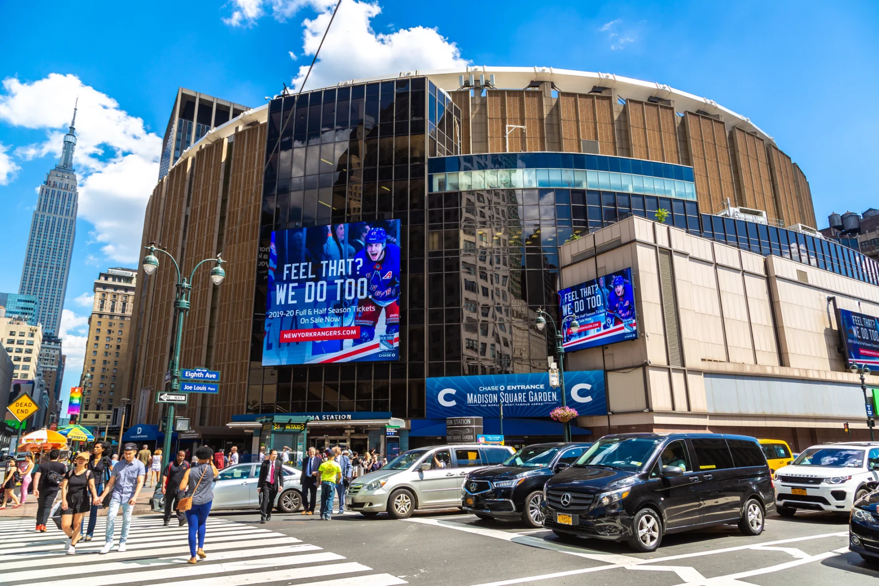 Außenansicht des Madison Square Garden in New York City, mit Menschen, die die Straße überqueren, und Autos im Straßenverkehr an einem sonnigen Tag. Im Hintergrund ist das Empire State Building zu sehen, ein Ort, an dem Stars wie Taylor Swift bereits aufgetreten sind. Alt-Text wurde KI-generiert.