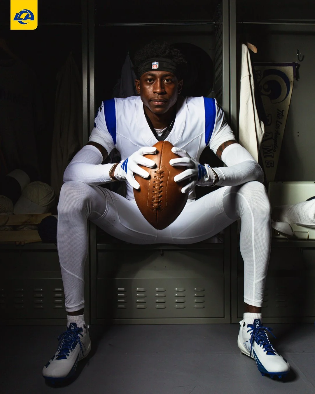 Ein Footballspieler in einem weiß-blauen Trikot – dem neuen Trikot der Los Angeles Rams – sitzt in einer Umkleidekabine, hält einen Football mit beiden Händen fest und blickt direkt in die Kamera. Alt-Text wurde KI-generiert.