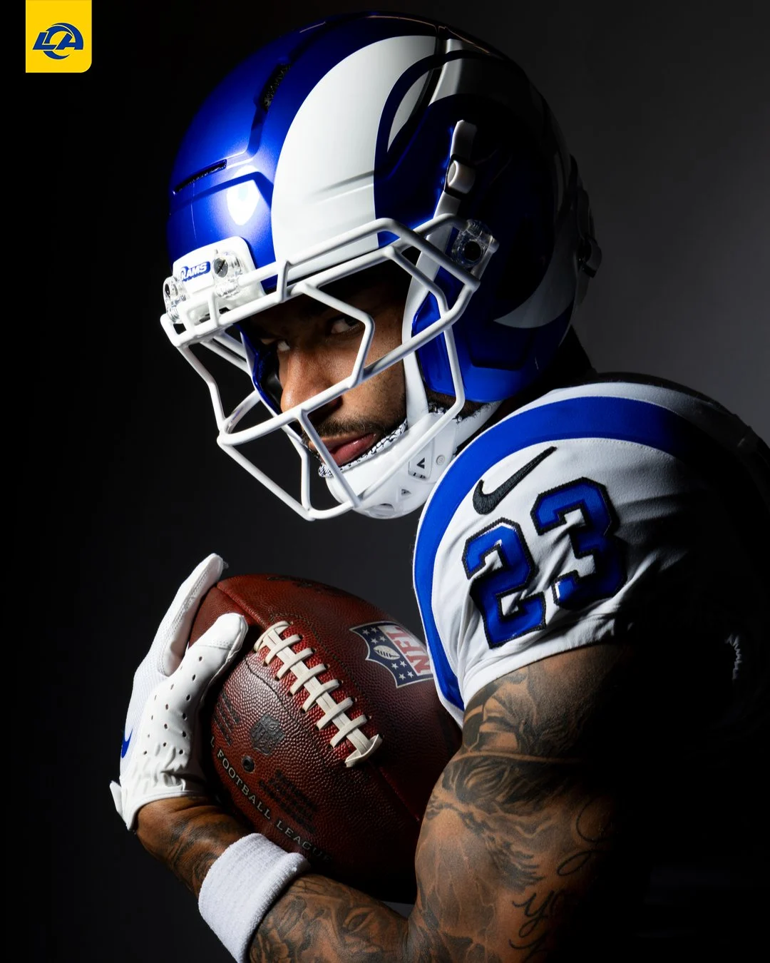 Ein Footballspieler im Trikot und Helm der Los Angeles Rams hält einen Football fest an sich gedrückt und trägt stolz das neue Trikot des Teams mit der Rückennummer 23. Alt-Text wurde KI-generiert.