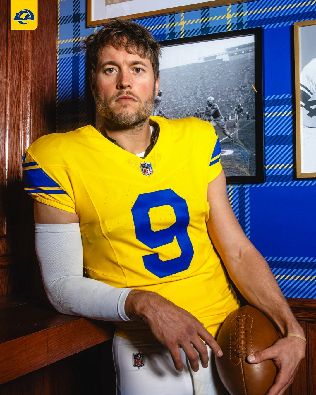 Ein Footballspieler im gelb-blauen Trikot der Los Angeles Rams hält stolz einen Football in der Hand, während er in einem Raum vor gerahmten Sportfotos an einer blau karierten Wand steht und die neuen Trikots des Teams präsentiert. Alt-Text wurde KI-generiert.
