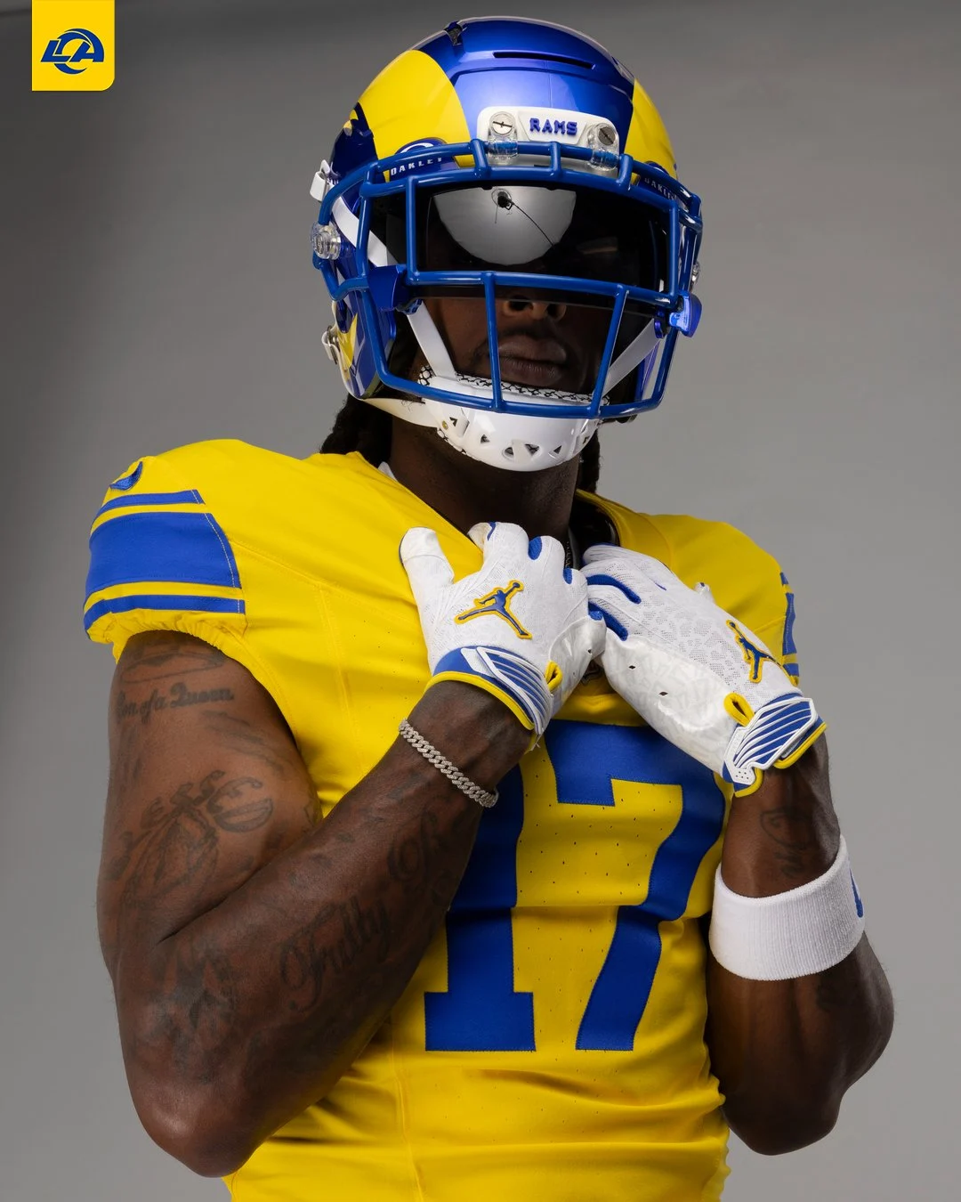 Ein NFL-Spieler in einem gelb-blauen Trikot und Helm der Los Angeles Rams posiert mit vor der Brust gefalteten Händen in Handschuhen, zeigt seine Tattoos auf den Armen und trägt stolz die neuen Trikots des Teams. Alt-Text wurde KI-generiert.