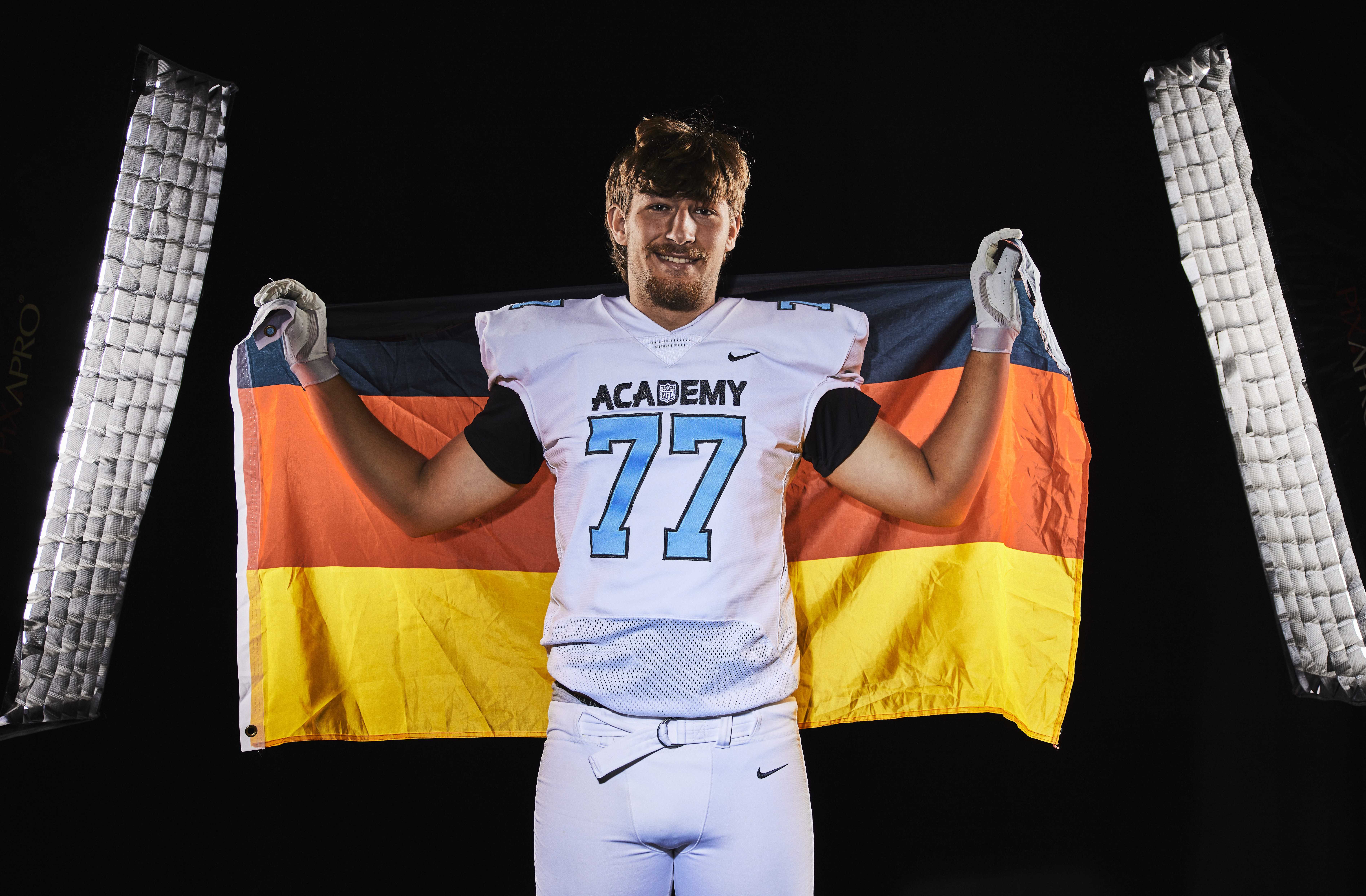 Ein Footballspieler in einem weißen „ACADEMY 77“-Trikot hält hinter sich eine deutsche Flagge und steht zwischen zwei senkrechten Lichtern vor einem schwarzen Hintergrund – O-Line-Talent Niko Kampas präsentiert stolz sein Können und repräsentiert damit sowohl Tennessee als auch seine Herkunft. Alt-Text wurde KI-generiert.