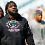 Ein Mann, der einen schwarzen Kapuzenpulli mit der Aufschrift „Crucial Catch Intercept Cancer“ und einem Football-Logo trägt, steht im Freien, während im Hintergrund mehrere Personen unscharf zu sehen sind – eine Szene, die an Packers-Defensive-Tackle Devonte Wyatt nach der Unterzeichnung seines 57-Millionen-Vertrags erinnert. Alt-Text wurde KI-generiert.