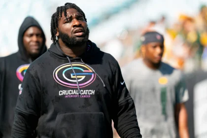Ein Mann, der einen schwarzen Kapuzenpulli mit der Aufschrift „Crucial Catch Intercept Cancer“ und einem Football-Logo trägt, steht im Freien, während im Hintergrund mehrere Personen unscharf zu sehen sind – eine Szene, die an Packers-Defensive-Tackle Devonte Wyatt nach der Unterzeichnung seines 57-Millionen-Vertrags erinnert. Alt-Text wurde KI-generiert.