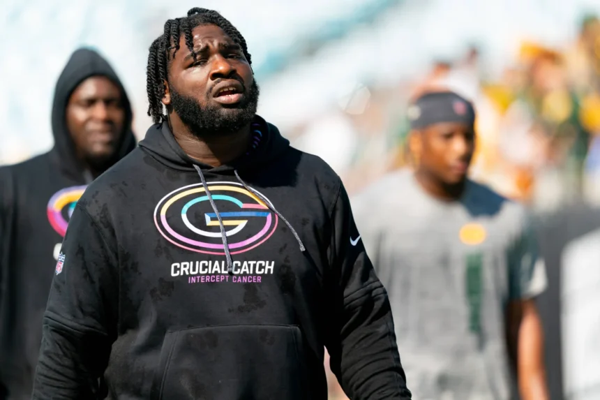 Ein Mann, der einen schwarzen Kapuzenpulli mit der Aufschrift „Crucial Catch Intercept Cancer“ und einem Football-Logo trägt, steht im Freien, während im Hintergrund mehrere Personen unscharf zu sehen sind – eine Szene, die an Packers-Defensive-Tackle Devonte Wyatt nach der Unterzeichnung seines 57-Millionen-Vertrags erinnert. Alt-Text wurde KI-generiert.
