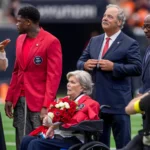 Eine Frau im Rollstuhl, die Rosen in den Händen hält und als Janice McNair identifiziert wurde, ist auf dem Footballfeld während einer Feier der Houston Texans zu sehen, umgeben von Menschen in Anzügen und Jacken. Alt-Text wurde KI-generiert.