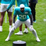 Ein Footballspieler im blaugrünen Trikot mit der Nummer 20 trainiert auf einem Rasenfeld, beugt sich in einer Bereitschaftsstellung nach vorne, während seine Teamkollegen in der Nähe stehen. Die Dolphins bereiten sich nach der Vertragsverlängerung von Jordyn Brooks mit neuer Energie auf die Saison vor. Alt-Text wurde KI-generiert.