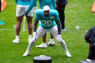 Ein Footballspieler im blaugrünen Trikot mit der Nummer 20 trainiert auf einem Rasenfeld, beugt sich in einer Bereitschaftsstellung nach vorne, während seine Teamkollegen in der Nähe stehen. Die Dolphins bereiten sich nach der Vertragsverlängerung von Jordyn Brooks mit neuer Energie auf die Saison vor. Alt-Text wurde KI-generiert.