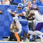 Ein Footballspieler in einem blauen Kentucky-Trikot springt in die Endzone, während er von einem gegnerischen Spieler in einem weiß-lila Trikot verfolgt wird – eine Szene, die an Georgias Verteidiger erinnert, die Dante Dowdell über das Spielfeld jagen. Im Hintergrund sind Fotografen und Zuschauer zu sehen. Alt-Text wurde KI-generiert.