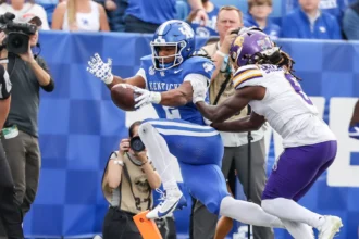 Ein Footballspieler in einem blauen Kentucky-Trikot springt in die Endzone, während er von einem gegnerischen Spieler in einem weiß-lila Trikot verfolgt wird – eine Szene, die an Georgias Verteidiger erinnert, die Dante Dowdell über das Spielfeld jagen. Im Hintergrund sind Fotografen und Zuschauer zu sehen. Alt-Text wurde KI-generiert.