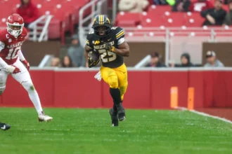 Der Footballspieler Ahmad Hardy aus Missouri läuft während eines Spiels gegen Arkansas mit dem Ball über den regennassen Rasen, während sich von links ein Verteidiger nähert – und beweist damit seine Widerstandsfähigkeit, obwohl er sich erst kürzlich von einer Schussverletzung erholt hat. Alt-Text wurde KI-generiert.