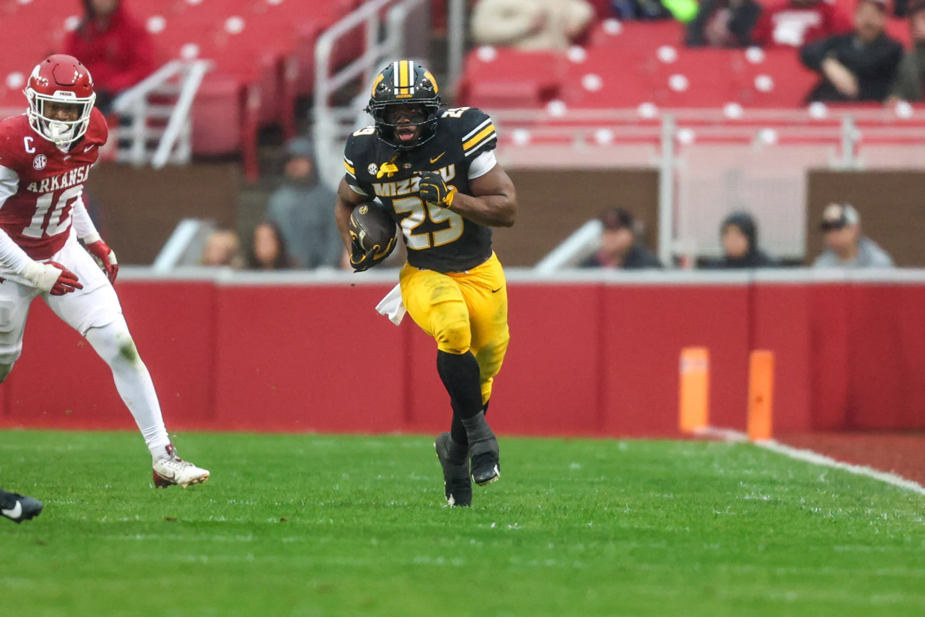 Der Footballspieler Ahmad Hardy aus Missouri läuft während eines Spiels gegen Arkansas mit dem Ball über den regennassen Rasen, während sich von links ein Verteidiger nähert – und beweist damit seine Widerstandsfähigkeit, obwohl er sich erst kürzlich von einer Schussverletzung erholt hat. Alt-Text wurde KI-generiert.