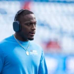 Nic Scourton in einem blauen Langarmhemd, mit Kopfhörern und einer Halskette steht in einem Stadion, im Hintergrund sind leere blaue Sitzplätze zu sehen, und lässt die Atmosphäre auf sich wirken, die an klassische „Panthers-Drama“-Momente erinnert. Alt-Text wurde KI-generiert.