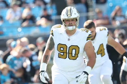 Ein Footballspieler in einem weißen Trikot der New Orleans Saints mit der Rückennummer 90, Bryan Bresee, steht während eines Spiels auf dem Spielfeld; im Hintergrund sind Teamkollegen und Fans zu sehen. Alt-Text wurde KI-generiert.