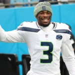 Nick Emmanwori, ein Footballspieler in einem weißen Trikot der Seattle Seahawks und einer Strickmütze, lächelt und winkt in der ersten Spielwoche am Spielfeldrand eines Footballfeldes. Alt-Text wurde KI-generiert.