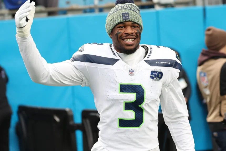 Nick Emmanwori, ein Footballspieler in einem weißen Trikot der Seattle Seahawks und einer Strickmütze, lächelt und winkt in der ersten Spielwoche am Spielfeldrand eines Footballfeldes. Alt-Text wurde KI-generiert.