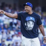 Ein Mann mit einer LA-Dodgers-Kappe und einem T-Shirt mit einem aufgedruckten Gesicht, der auf einem Baseballfeld nach links zeigt, im Hintergrund die Zuschauermenge – ein Bild, das die Aufregung eines Spieltags einfängt und an Rams-Fans erinnert, die Myles Garrett bei den „Camp-Tag“-Veranstaltungen entdecken. Alt-Text wurde KI-generiert.