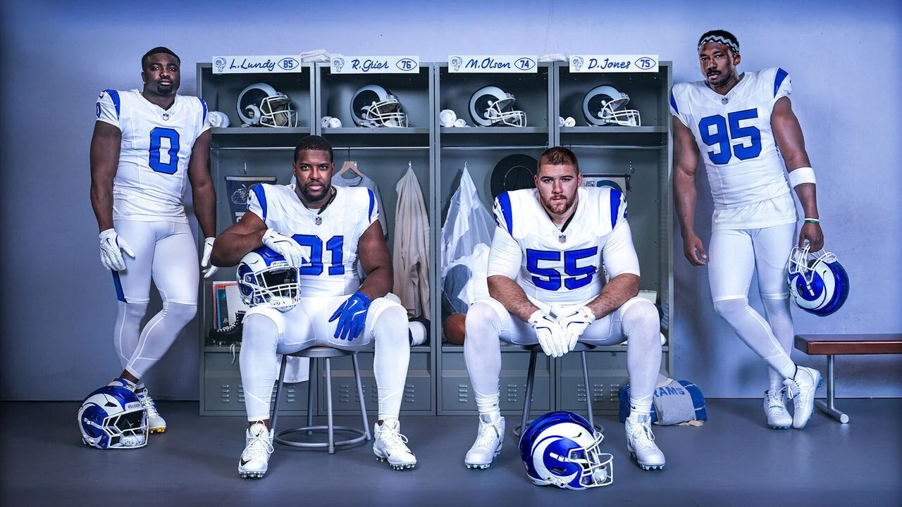 Vier Footballspieler der Los Angeles Rams in weiß-blauen Trikots posieren vor ihren Spinden, während ihre Helme auf dem Boden liegen und ihre Namen über den Spinden zu sehen sind. Alt-Text wurde KI-generiert.
