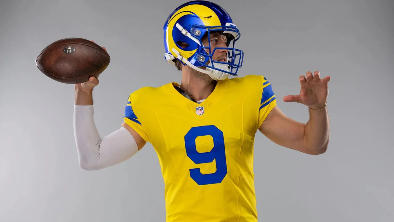 Matthew Stafford in einem gelb-blauen Trikot mit der Nummer 9 macht sich bereit, einen Football zu werfen. Er trägt einen Helm, blickt nach links und präsentiert stolz die neuen Trikots der Los Angeles Rams. Alt-Text wurde KI-generiert.