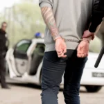 Ein Polizeibeamter legt einem Mann, der sich als 49ers Spieler ausgibt und Tätowierungen auf dem Rücken hat, in der Nähe eines Streifenwagens Handschellen an, während im Hintergrund ein weiterer Beamter steht. Alt-Text wurde KI-generiert.