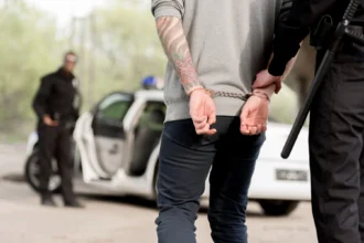 Ein Polizeibeamter legt einem Mann, der sich als 49ers Spieler ausgibt und Tätowierungen auf dem Rücken hat, in der Nähe eines Streifenwagens Handschellen an, während im Hintergrund ein weiterer Beamter steht. Alt-Text wurde KI-generiert.