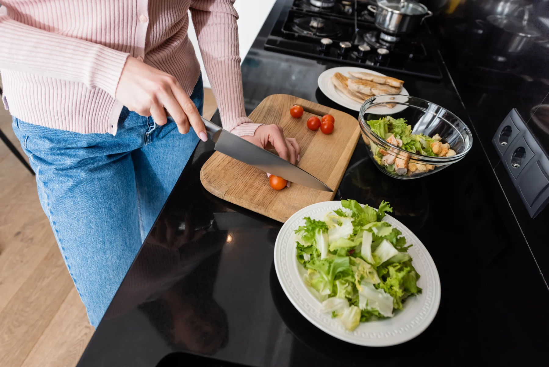 Eine Person schneidet auf einem schwarzen Küchentisch neben Tellern mit Salat und gegartem Hähnchen auf einem Holzschneidebrett Kirschtomaten in Scheiben und bereitet „High-Protein Mediterrane Kartoffeln mit Hähnchen“ als köstliche und ausgewogene Mahlzeit zu. Alt-Text wurde KI-generiert.