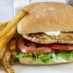 Sandwich mit gegrilltem Peri-Peri-Hähnchen und Halloumi auf Ciabatta aus dem Airfryer, mit Salat, Tomate und Mayonnaise auf einem Ciabatta-Brötchen, serviert mit gewürzten Pommes frites auf einem weißen Teller. Alt-Text wurde KI-generiert.