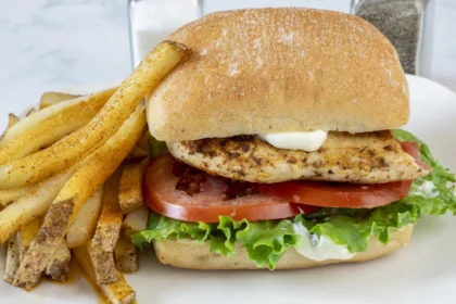 Sandwich mit gegrilltem Peri-Peri-Hähnchen und Halloumi auf Ciabatta aus dem Airfryer, mit Salat, Tomate und Mayonnaise auf einem Ciabatta-Brötchen, serviert mit gewürzten Pommes frites auf einem weißen Teller. Alt-Text wurde KI-generiert.