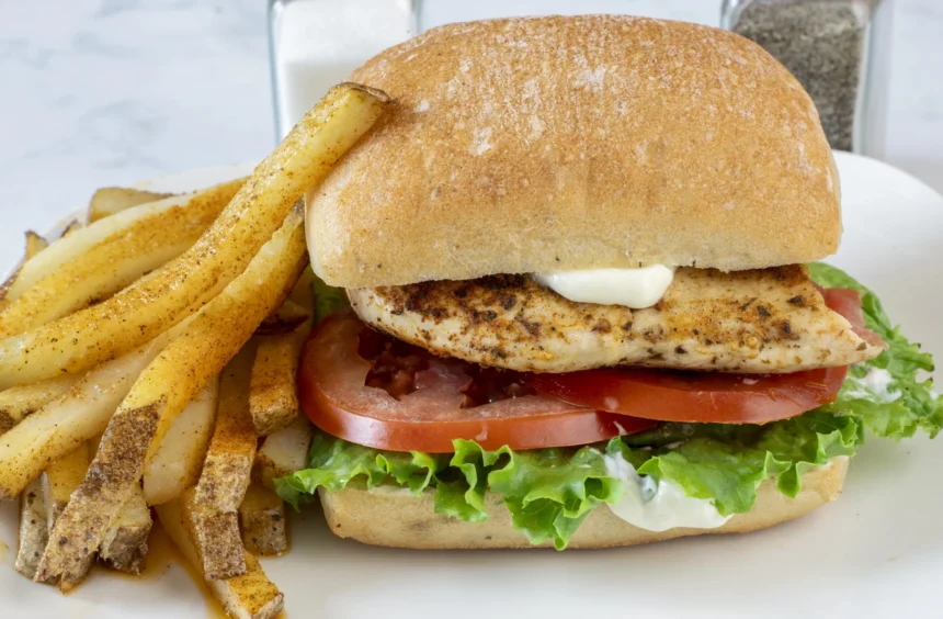 Sandwich mit gegrilltem Peri-Peri-Hähnchen und Halloumi auf Ciabatta aus dem Airfryer, mit Salat, Tomate und Mayonnaise auf einem Ciabatta-Brötchen, serviert mit gewürzten Pommes frites auf einem weißen Teller. Alt-Text wurde KI-generiert.