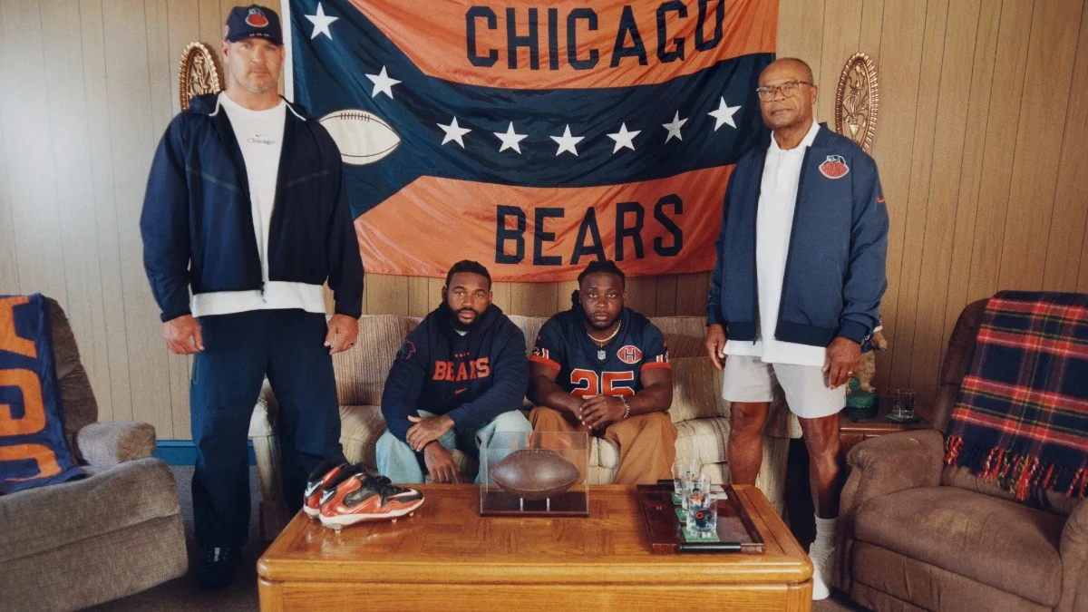 Vier Männer in Chicago-Bears-Outfits posieren in einem Wohnzimmer vor Bears-Fanartikeln, darunter eine große Flagge, ein Football und Team-Ausrüstung auf einem hölzernen Couchtisch. Sie tragen Kleidung aus der „NFL RivalriesKollektion“ von Nike, die sowohl ihre Teamtreue als auch ihren Sinn für Stil unterstreicht. Alt-Text wurde KI-generiert.