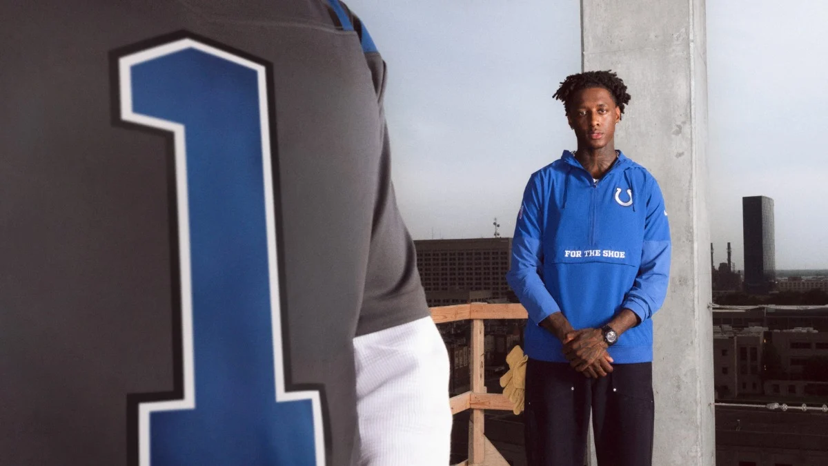 Ein Mann in einem blauen Colts-Hoodie steht auf einem Balkon mit Blick auf die Stadt; im Vordergrund ist eine Person in einem grauen NFL-Trikot mit einer großen blauen „1“ zu sehen, was auf ihre Leidenschaft für klassische Rivalitäten und exklusive Stücke wie die RivalriesKollektion hindeutet. Alt-Text wurde KI-generiert.