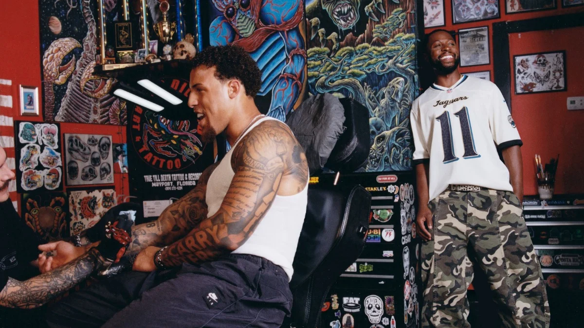Zwei Männer in einem Tattoo-Studio: Der eine lässt sich auf einem Stuhl sitzend ein Tattoo auf den Unterarm stechen, der andere steht lächelnd daneben. Im Hintergrund sind farbenfrohe Kunstwerke und Trophäen zu sehen, dazu Nike-Ausrüstung aus der exklusiven „RivalriesKollektion“ für NFL-Fans. Alt-Text wurde KI-generiert.